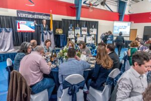 Anne Gore talks at the podium as guests mingle and eat meals provided by Bridge Seafood Restaurent at Lumen Christi Catholic High Schools’ 28th Annual Fundraising Gala on Feb. 11, 2023.